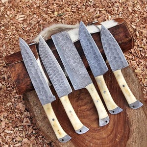 May include: Five Damascus steel knives with bone handles are displayed on a wooden surface. The blades have intricate patterns, and the handles are a creamy ivory color. A leather case with a cowhide flap is visible in the background.