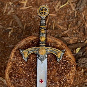 Op de afbeelding: Een decoratief zwaard met een rijk versierd gevest met goud- en zilverkleurige metalen details, een ronde pommel met een kruisembleem en een lemmet gegraveerd met ingewikkeld krulwerk en een rood kruissymbool bij de stootplaat.