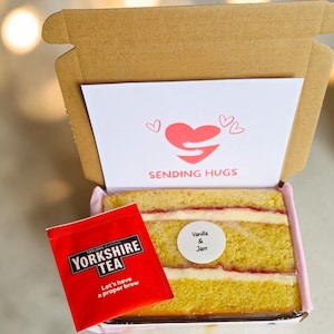 May include: A brown cardboard gift box containing a slice of vanilla and jam cake and a red packet of Yorkshire Tea. The box lid features a white card with a red heart graphic and the text SENDING HUGS. A round sticker on the cake reads Vanilla & Jam.