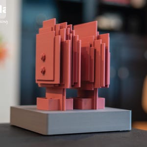 May include: A coral-colored, geometric sculpture with a layered design, mounted on a gray rectangular base. The sculpture has a pixelated appearance and is set against a blurred background. The Della logo and the text "The art of making" are in the upper left corner.
