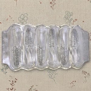 May include: A clear glass serving dish featuring six moulded indentations, each shaped like an ear of corn. The rectangular tray has scalloped edges and a smooth, transparent finish, designed to hold individual corn cobs for serving.