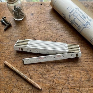 May include: A white and gold folding ruler with black markings, measuring up to 12 inches. A wooden pencil with text and a blueprint are also visible. A small glass jar with screws and bolts is in the background. The ruler is partially open.