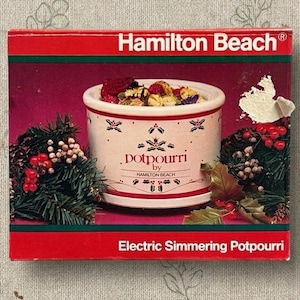 May include: A Hamilton Beach electric simmering potpourri box featuring a white ceramic pot with red holly leaf patterns and text that reads potpourri by Hamilton Beach. The box displays a festive arrangement of pine branches and red berries.