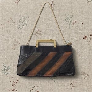 May include: A rectangular handbag featuring a textured black and brown diagonal striped pattern. The bag includes a rigid gold-tone metal top handle and a thin gold-tone metal chain shoulder strap attached to the sides of the bag's upper edge.