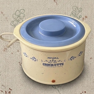May include: A cream-coloured Rival Crock-Ette stoneware slow cooker with a speckled finish and blue floral accents. It features a round blue lid, a side handle, and a power cord. The front displays the text RIVAL CROCK-ETTE STONEWARE SLOW COOKER.