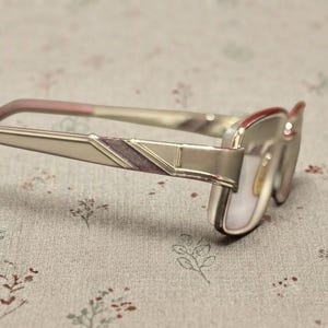 May include: A pair of spectacles featuring a metallic champagne-coloured frame with a rectangular shape. The temples are accented with a diagonal, textured plum-coloured inlay. The bridge and hinges are integrated into the sleek, structured metal design.