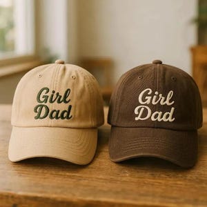May include: Two baseball caps are shown, one beige and one brown. The beige cap has green embroidered text reading "Girl Dad." The brown cap has white embroidered text reading "Girl Dad."