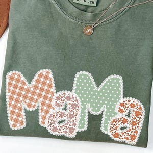 May include: An olive green Comfort Colors t-shirt with the word "Mama" in a patchwork design. The "M" is a brown plaid, the first "a" is a floral print, the second "m" is white polka dots, and the second "a" is a floral print. A gold necklace rests on the shirt.