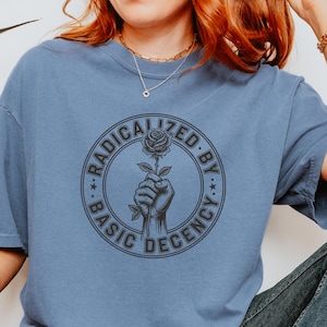 以下が含まれることがあります： 「RADICALIZED BY BASIC DECENCY」の文字が円形デザインでプリントされた青いTシャツ。デザインはバラを持つ手を描いています。Tシャツは無地の青色で、柔らかい素材で作られています。