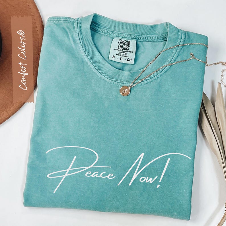 May include: A teal Comfort Colors t-shirt with the words "Peace Now!" in white script. A gold necklace with a pendant rests on the shirt. The shirt is folded neatly, and the tag is visible. A brown hat is in the background.