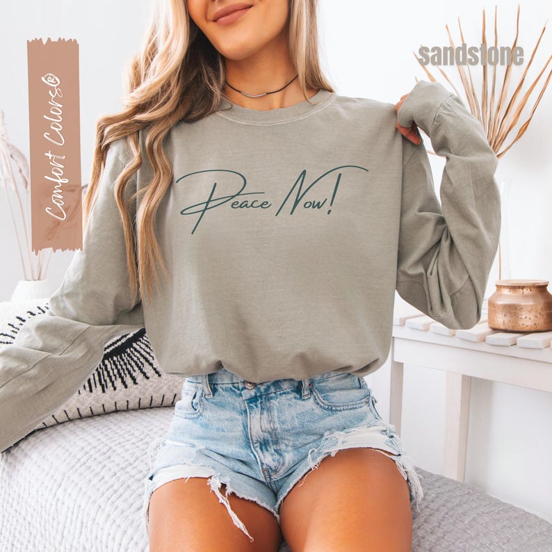 May include: A long-sleeved, sage green sweatshirt with the words "Peace Now!" in a cursive font. The sweatshirt is paired with denim shorts. The image also includes the text "Comfort Colors" and "sandstone".