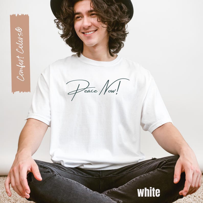 May include: White t-shirt with the text "Peace Now!" in a cursive font. The word "white" is at the bottom of the shirt. The shirt is part of the "Comfort Colors" collection.