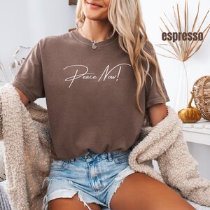 May include: Espresso-colored t-shirt with the words "Peace Now!" in white script. The shirt is a short-sleeved, crew neck style. The model is wearing denim shorts and a cream-colored cardigan.
