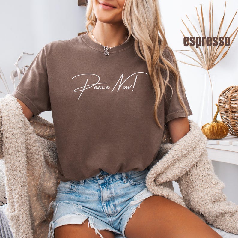 May include: Espresso-colored t-shirt with the words "Peace Now!" in white script. The shirt is a short-sleeved, crew neck style. The model is wearing denim shorts and a cream-colored cardigan.