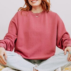 Op de afbeelding: Een stoffig roze sweatshirt met lange mouwen. Het sweatshirt wordt gecombineerd met lichtblauwe jeans met distressed details. Een zilveren ketting is zichtbaar. De persoon glimlacht.