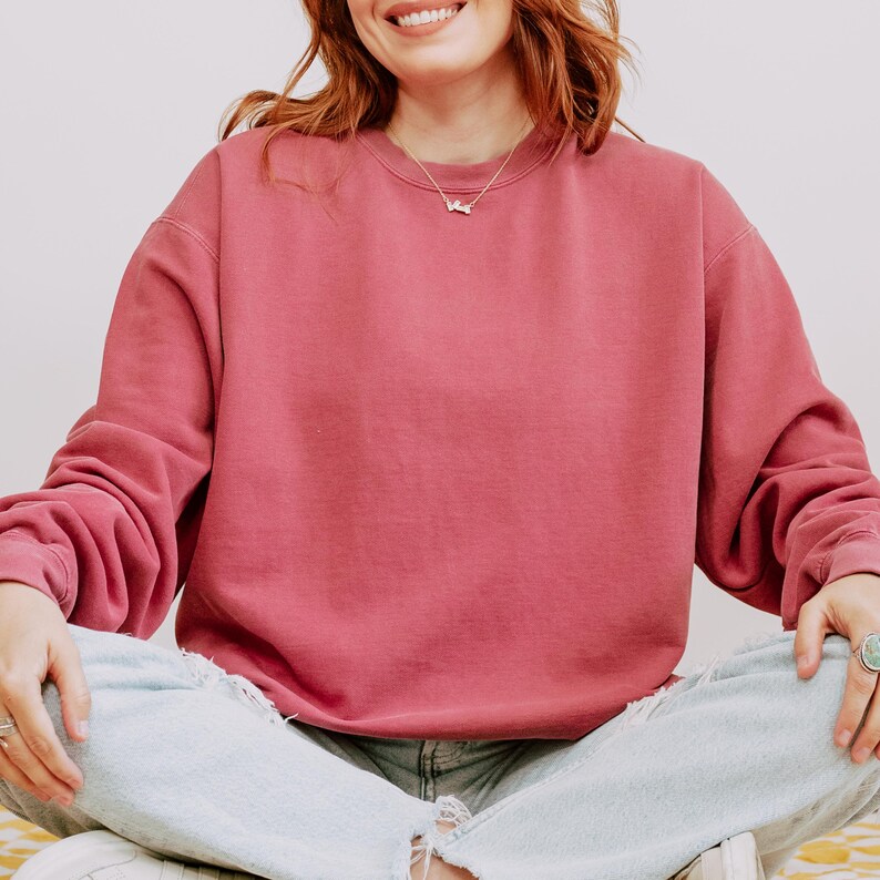 Op de afbeelding: Een stoffig roze sweatshirt met lange mouwen. Het sweatshirt wordt gecombineerd met lichtblauwe jeans met distressed details. Een zilveren ketting is zichtbaar. De persoon glimlacht.