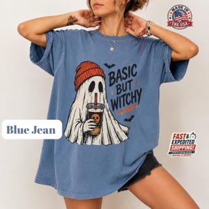 May include: A blue jean colored t-shirt featuring a graphic of a white ghost wearing an orange beanie and holding a coffee cup. The text "BASIC BUT WITCHY" is printed in black next to the ghost, accompanied by small black bat silhouettes.