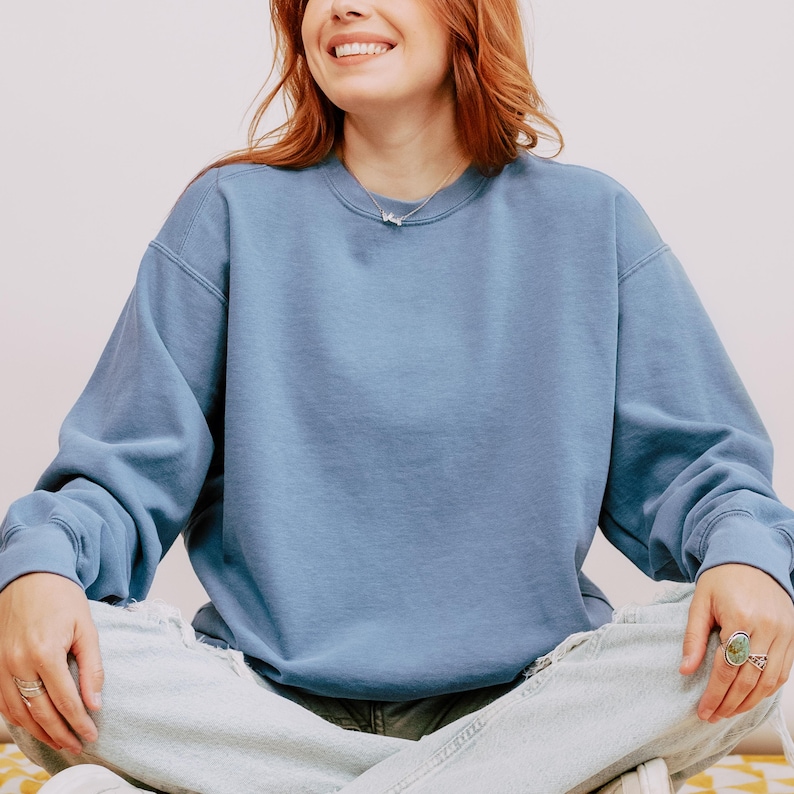 Op de afbeelding: Een lichtblauw sweatshirt met een ronde hals. De top met lange mouwen wordt gedragen met een lichtgebleekte jeans. De persoon draagt ringen en een zilveren ketting. De achtergrond is licht.
