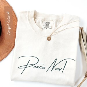 May include: Cream-colored Comfort Colors t-shirt with the words "Peace Now!" in a dark teal script. A gold necklace with a pendant rests on the shirt. The shirt is folded neatly on a white surface.