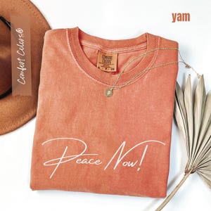 May include: A yam-colored Comfort Colors t-shirt with the words "Peace Now!" in white script. A gold necklace with a pendant rests on the shirt. A brown hat and dried palm leaf are in the background.