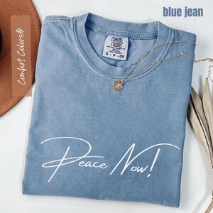 May include: A blue jean colored t-shirt with the words "Peace Now!" in white script. A gold necklace with a pendant rests on the shirt. The shirt has a Comfort Colors tag.