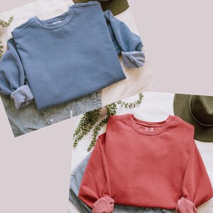 Op de afbeelding: Twee crewneck sweatshirts. E&eacute;n is stofblauw, de andere is koraal. Beide hebben opgerolde mouwen. Ze worden gepresenteerd op een wit oppervlak met een spijkerbroek en een groene hoed. De sweatshirts zijn gemaakt van een zacht materiaal.