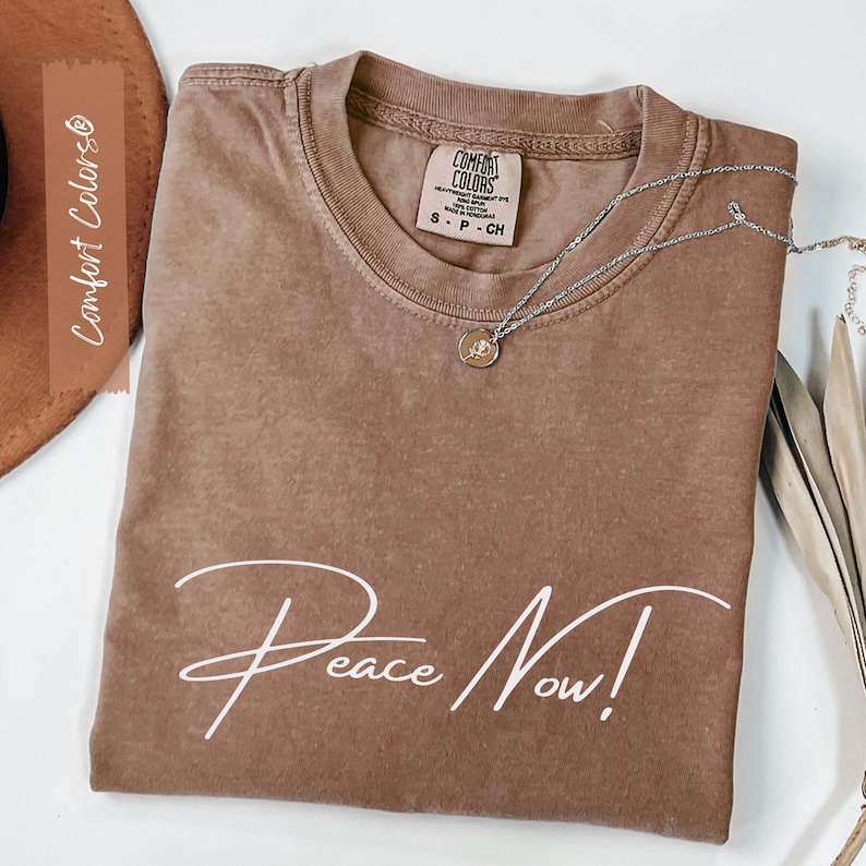 May include: A brown Comfort Colors t-shirt with the words "Peace Now!" in white script. A silver necklace with a small pendant rests on the shirt. The shirt is folded and displayed with a brown hat and dried leaves.