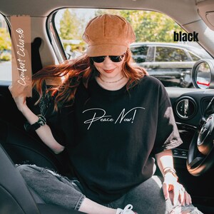 May include: A black t-shirt with the words "Peace Now!" in white script. The person in the image is wearing a brown corduroy cap and sunglasses. The image is taken inside a car.