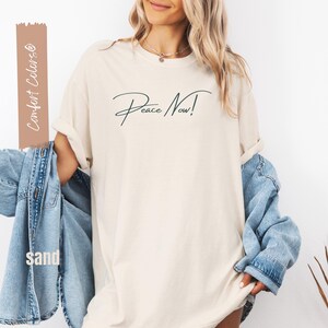 May include: A sand-colored t-shirt with the words "Peace Now!" in a dark green script. The shirt is paired with a light blue denim jacket. The words "Comfort Colors" and "sand" are printed on a vertical tan rectangle.