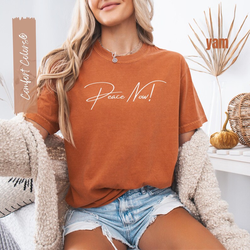 May include: A yam-colored t-shirt with the words "Peace Now!" in a white script. The shirt is paired with denim shorts and a cream-colored cardigan. The words "Comfort Colors" are printed on a vertical brown rectangle.