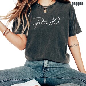 May include: Dark gray t-shirt with the words "Peace Now!" in white script. The shirt is a relaxed fit and is paired with blue jeans. The word "pepper" is printed on the top right corner of the shirt.