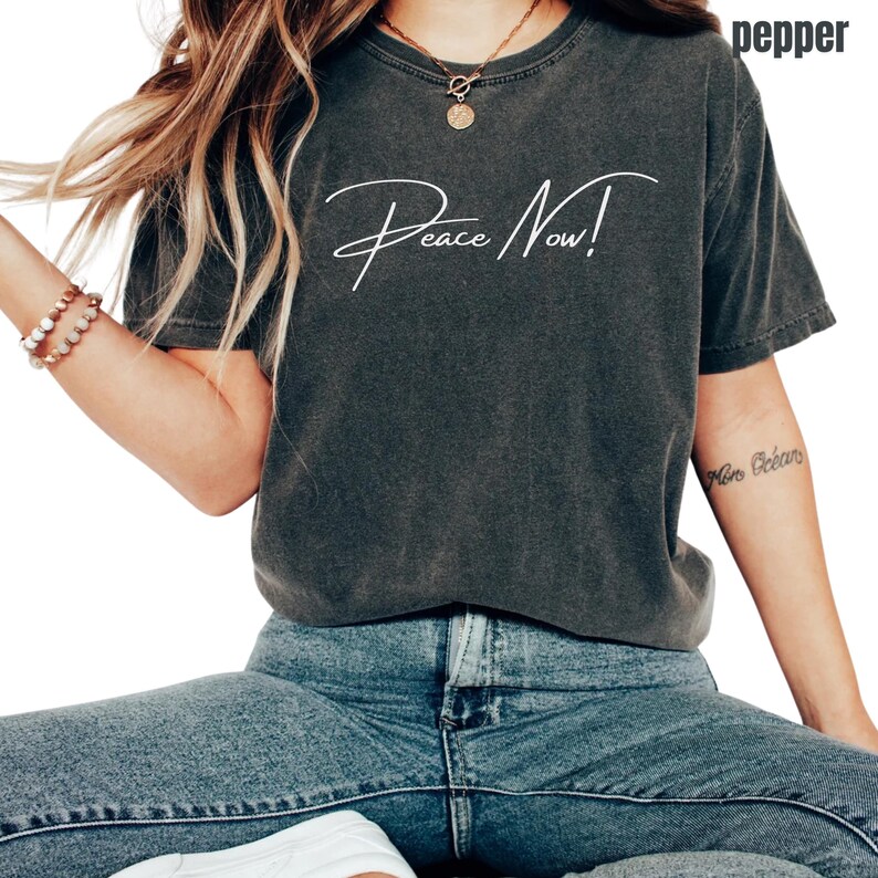 May include: Dark gray t-shirt with the words "Peace Now!" in white script. The shirt is a relaxed fit and is paired with blue jeans. The word "pepper" is printed on the top right corner of the shirt.