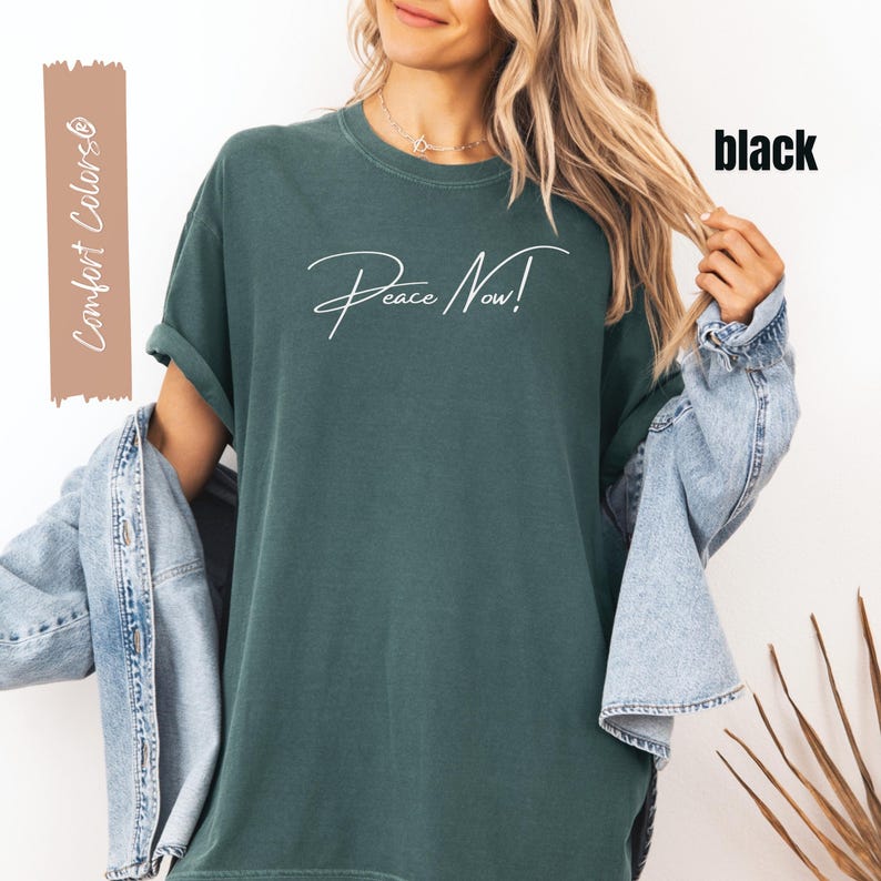 May include: Dark green t-shirt with the text "Peace Now!" in white script. The shirt has rolled-up sleeves and is paired with a denim jacket. The words "Comfort Colors" are printed on a brown rectangle.