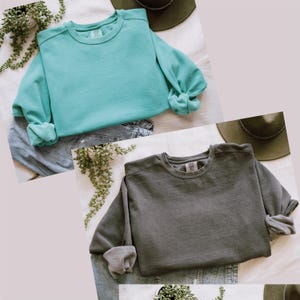 Op de afbeelding: Twee crewneck sweatshirts, &eacute;&eacute;n in teal en &eacute;&eacute;n in grijs, met geknoopte mouwen. De sweatshirts worden getoond op een spijkerbroek en een wit oppervlak. Een groene hoed en een plant staan ook in de afbeelding.