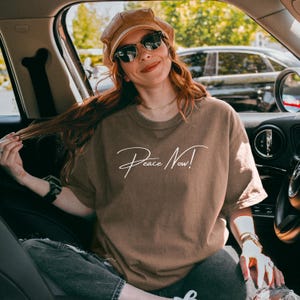 May include: A brown t-shirt with the words "Peace Now!" in white script. The person in the image is wearing sunglasses and a corduroy hat. The person is sitting in a car.