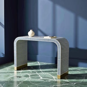 Op de afbeelding: Een moderne consoletafel met een gebogen ontwerp. De tafel heeft een wit en marineblauw gestreept patroon aan de zijkanten en bovenkant. De basis van de tafel heeft gouden accenten. Een kleine witte vaas en een boek staan op de tafel.