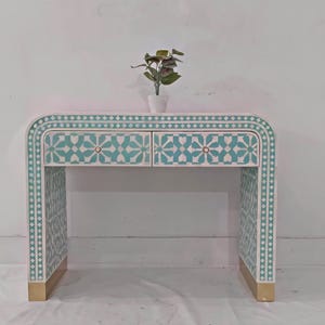 Puede incluir: Un escritorio blanco y turquesa con dos cajones y patas doradas. El escritorio tiene un diseño curvo y está decorado con un patrón geométrico. Una pequeña planta en maceta se encuentra encima del escritorio.