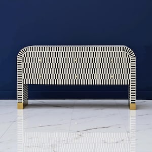 May include: A black and white patterned console table with a rectangular shape and rounded corners. The table has a repeating vertical stripe pattern and gold-colored feet. The background is a solid dark blue.
