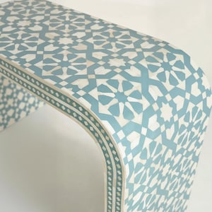 Op de afbeelding: Een gebogen tafel met een lichtblauw en wit geometrisch patroon. Het tafelblad heeft een herhalend ster- en ruitontwerp. De rand van de tafel heeft een decoratieve rand. De tafel is ongeveer 100 cm lang.