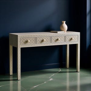 Könnte beinhalten: Eine cremefarbene Konsole mit drei Schubladen und einem geometrischen Muster. Auf dem Tisch stehen eine hellfarbige Vase und ein Buch. Die Schubladen haben runde Knöpfe. Der Tisch ist 122 cm lang.