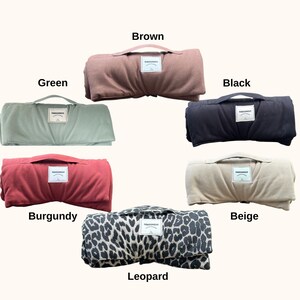 Op de afbeelding: Een verzameling opgerolde dekens in diverse kleuren: bruin, groen, zwart, bordeaux, beige en luipaardprint. Elke deken heeft een handvat en een klein label. Ontworpen voor gebruik buitenshuis.