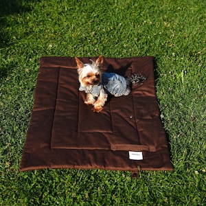 Pode incluir: Um cão pequeno usando uma jaqueta jeans descansa em uma cama para animais de estimação retangular marrom. A cama está sobre a grama. O cão tem pelo marrom e branco. A cama tem um design acolchoado e uma pequena etiqueta.