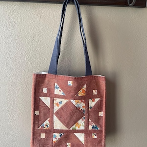 May include: A handmade tote bag with a patchwork design. The bag features a central diamond shape and star patterns, with floral fabric accents in the squares. The bag has a dark red-brown color and navy blue handles.