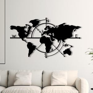 Puede incluir: Arte mural de mapa del mundo de metal negro con diseño de brújula. El mapa presenta continentes y está montado en una pared blanca sobre un sofá blanco. El diseño de la brújula incluye indicadores direccionales.