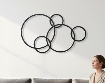 Pode incluir: Uma peça de arte de parede em metal preto composta por cinco anéis circulares interconectados de vários tamanhos. A escultura apresenta um design de moldura fina e contínua que se sobrepõe para criar um arranjo geométrico numa parede branca.