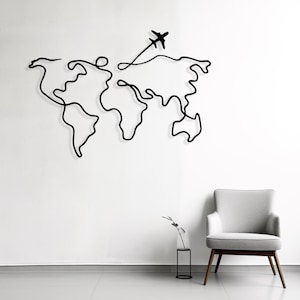 May include: Black metal wall art showing a continuous line drawing of a world map with an airplane. The artwork is mounted on a white wall, with a grey chair and minimalist decor in the background. A modern travel-themed home decor piece.