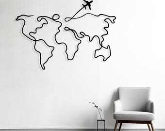 May include: Black metal wall art depicting a continuous line drawing of a world map with an airplane. The artwork is mounted on a white wall, with a gray chair and minimalist decor in the background. A modern travel-themed home decor piece.