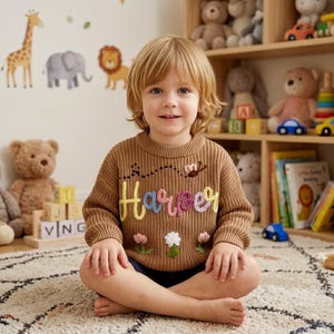 May include: A tan long-sleeve knit sweater featuring the name Harper embroidered in colorful yarn letters across the chest. The design includes a small stitched butterfly with a dotted flight path above the name and three small flowers below.