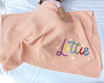 May include: A peach-coloured knitted baby blanket with the name "Lottie" embroidered in colourful thread. The blanket has a ribbed border and is decorated with small embroidered flowers.