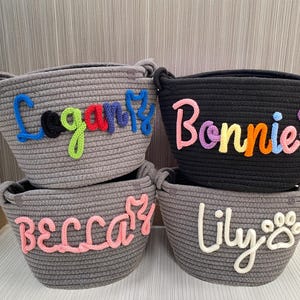May include: Four rope baskets in grey and black, each with a name embroidered on the front. The names are Logan, Bonnie, Becca, and Lily. The Lily basket has a paw print design.